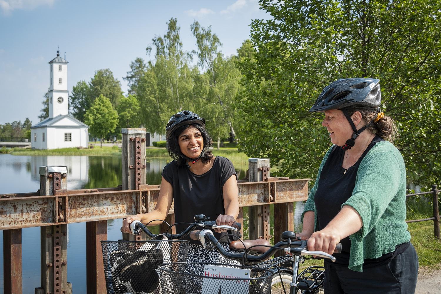 Zwei Frauen mit Fahrrädern stehen auf einer Brücke mit einer kleinen Kirche im Hintergrund.