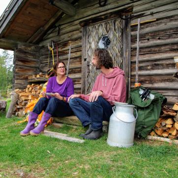Een man en een vrouw zitten voor een houten stuga met stapels hout ernaast.