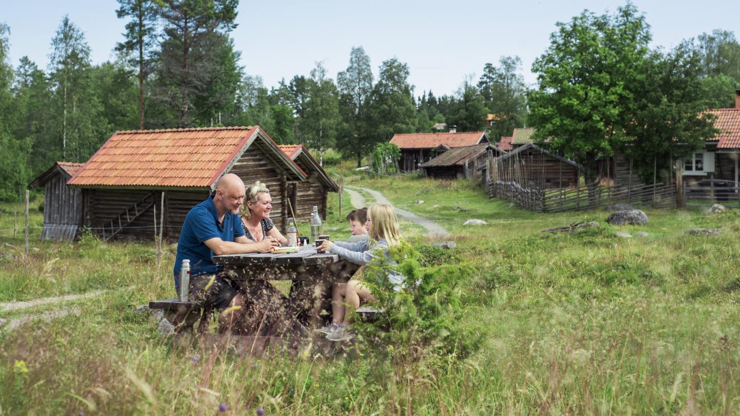 Twee volwassenen en twee kinderen picknicken tussen oude huisjes op een zomerboerderij.