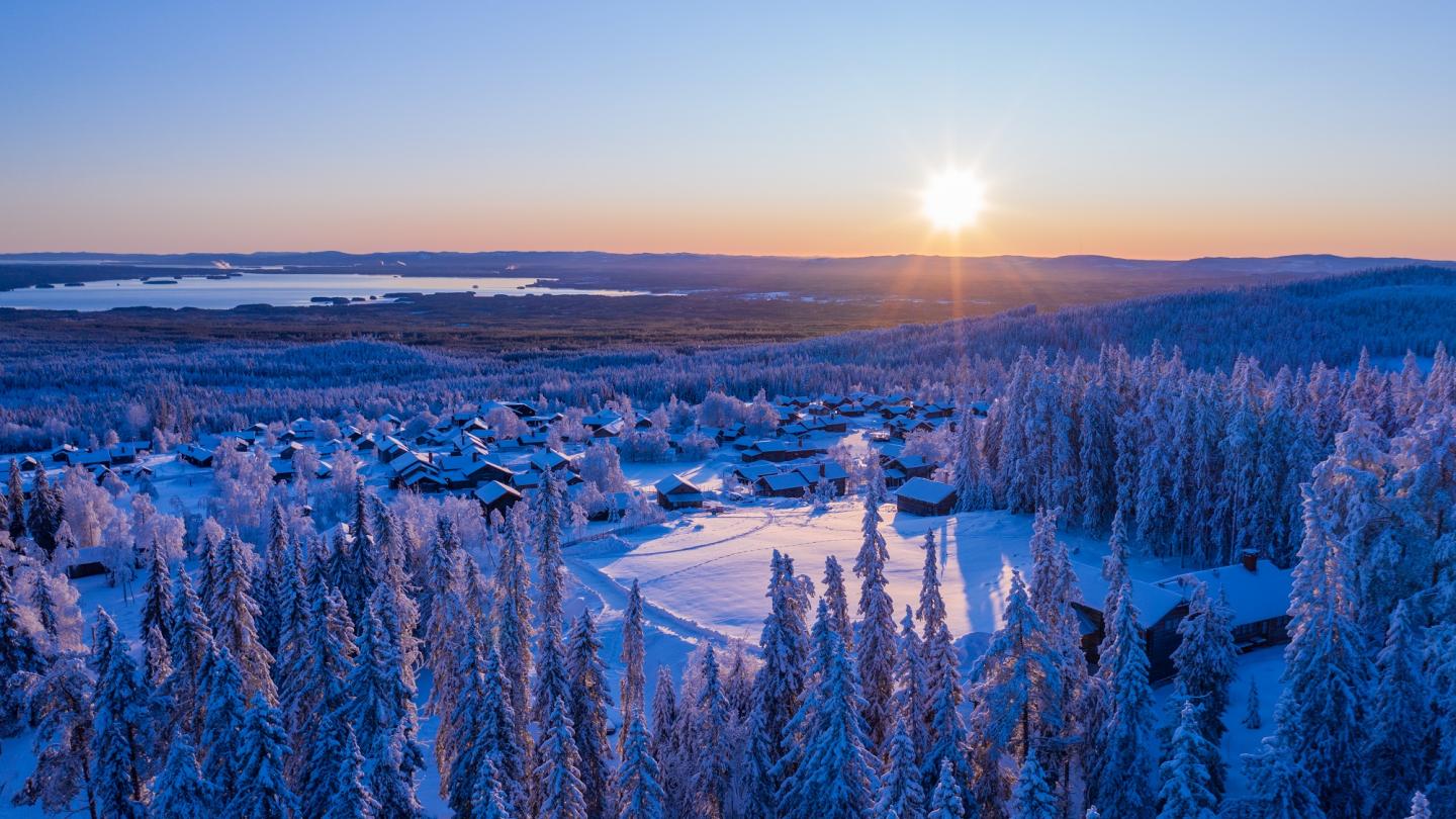 Eine untergehende Sonne in einer verschneiten Landschaft mit Hütten, Wald und See im Hintergrund.
